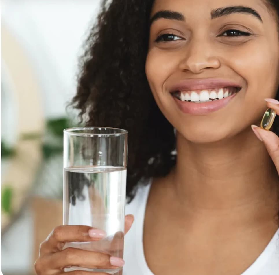Woman smiling, holding a glass of water and a vitamin capsule